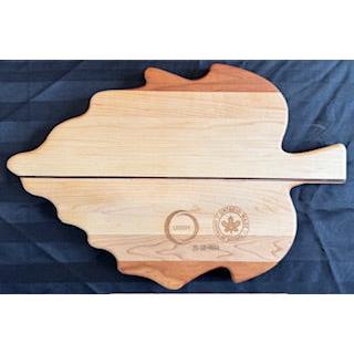 A wide leaf-shaped serving board with the words 'kitchen' and 'the heart of the home' engraved on it, with a combination of maple, walnut, and purple heart wood species.